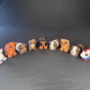 Peut inclure: Une collection de neuf figurines d'animaux comprenant un lapin gris, divers chiens marron et noirs, des cochons d'Inde et un poisson combattant blanc, rouge et bleu. Chaque miniature présente de grands yeux sombres et un aspect lisse.
