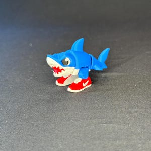 May include: A small, articulated blue shark figurine with a white underbelly and open mouth showing red teeth. The shark is wearing bright red sneakers with white soles and a small white swoosh logo on the side of each shoe, standing upright.