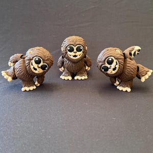 May include: Three brown, furry creature figurines with large black eyes and cream-colored teeth. The central figure stands upright, while the two flanking figures are posed in dynamic, leaning positions with one leg raised, showcasing textured fur.
