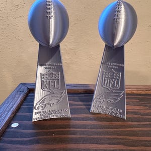 Pu&ograve; includere: Due trofei d'argento del Super Bowl NFL con il testo "Vince Lombardi Trophy" e il logo NFL. I trofei sono su una superficie di legno. Il testo sui trofei recita "Super Bowl LII Champions February 4, 2018".