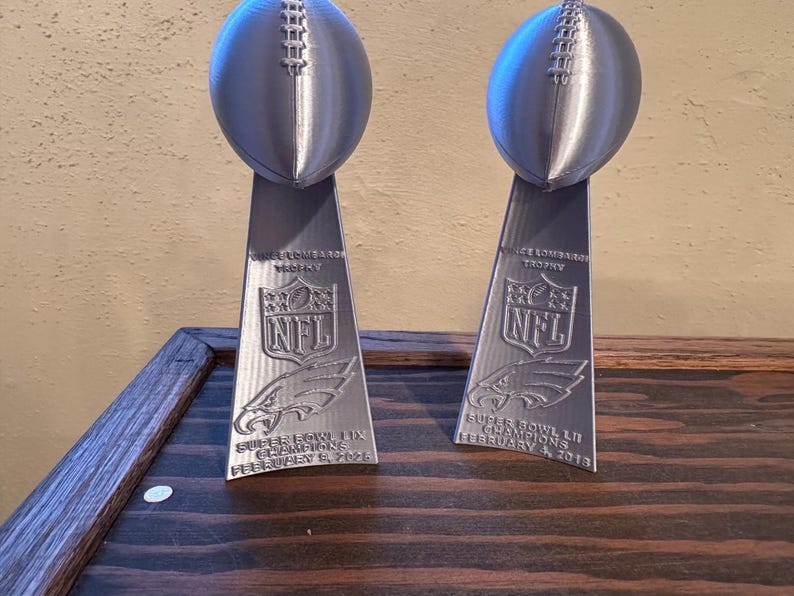 Pu&ograve; includere: Due trofei d'argento del Super Bowl NFL con il testo "Vince Lombardi Trophy" e il logo NFL. I trofei sono su una superficie di legno. Il testo sui trofei recita "Super Bowl LII Champions February 4, 2018".