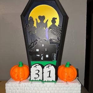 以下が含まれることがあります： ハロウィーンをテーマにした装飾で、棺型のフレームが特徴です。フレームには、魔女、家、満月のシルエットシーンが含まれています。2つのオレンジ色のカボチャと31の数字が入った墓石が前にあります。