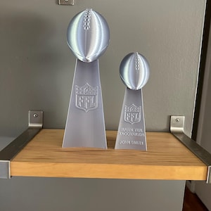 Pu&ograve; includere: Due trofei di football argentati su uno scaffale di legno. Il trofeo pi&ugrave; grande ha il logo NFL e il trofeo pi&ugrave; piccolo ha il testo "HAWK TUA TAGOVAILOA JOHN SMITH".
