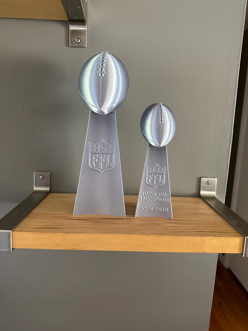 Pu&ograve; includere: Due trofei di football argentati su uno scaffale di legno. Il trofeo pi&ugrave; grande ha il logo NFL e il trofeo pi&ugrave; piccolo ha il testo "HAWK TUA TAGOVAILOA JOHN SMITH".