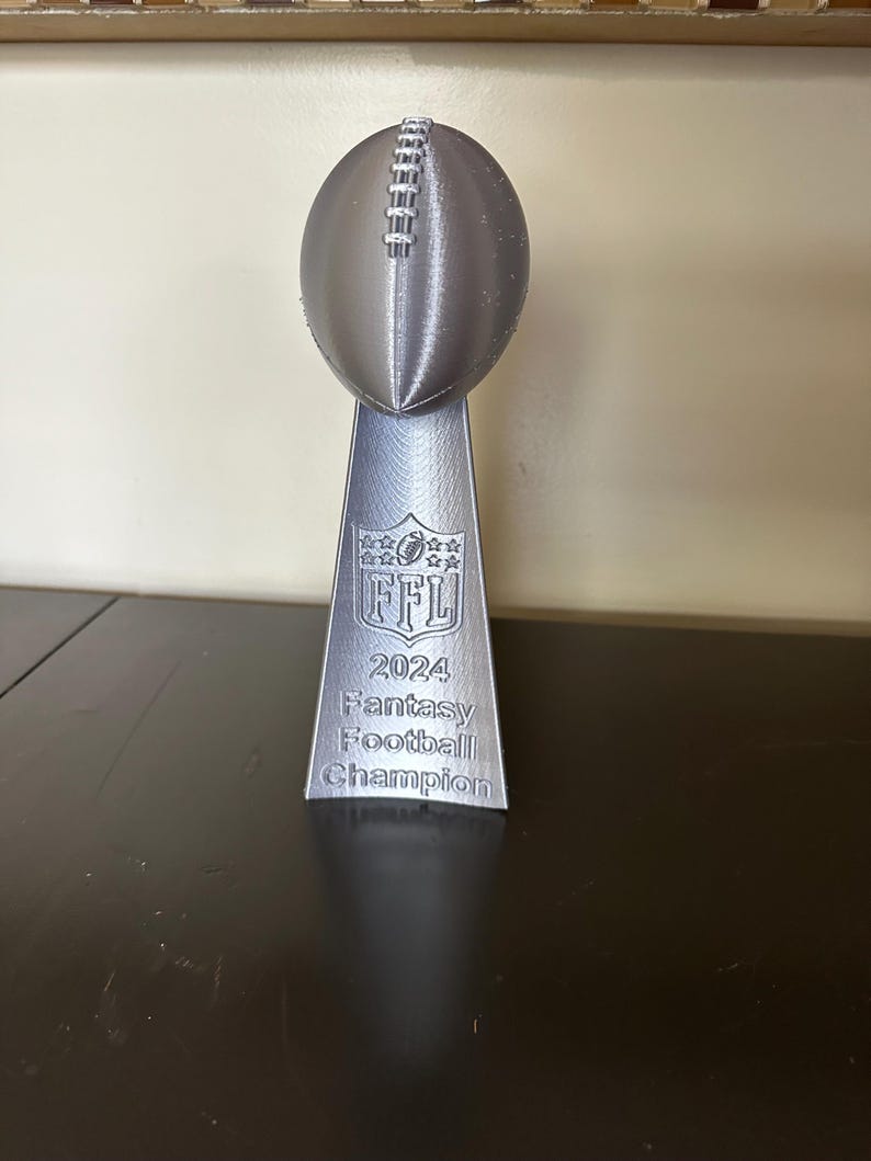 Pu&ograve; includere: Un trofeo argentato a forma di pallone da football con il testo "2024 Fantasy Football Champion" e il logo della NFL.