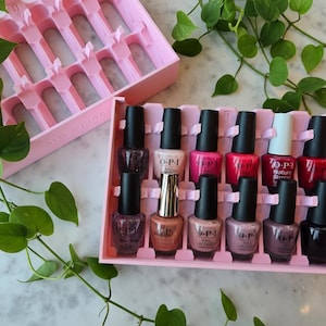 May include: Pink nail polish bottles with black caps are arranged in a pink organizer. The organizer is designed to hold the bottles securely. Various shades of nail polish are visible, including pinks, reds, and neutrals. The OPI brand is visible on some bottles.