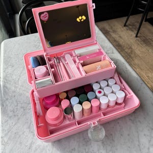 May include: A pink plastic nail supply organizer case with a built-in mirror featuring a heart-shaped sticker that says "OLIVE YOU". The case contains various nail tools, including brushes, files, buffers, and numerous small bottles of nail polish.