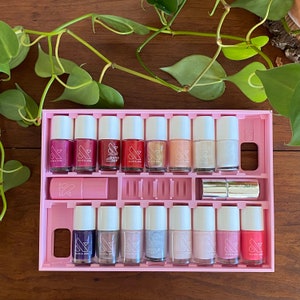 May include: A pink nail polish organiser with 18 clear bottles of nail polish. The bottles are arranged in two rows of nine. The bottles are labelled "Olive & June" and "Quick Dry".