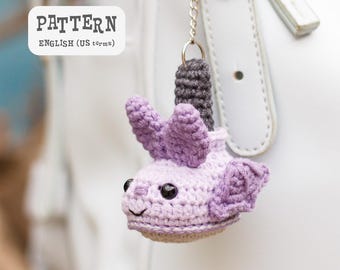 Puede incluir: Un llavero de ganchillo hecho a mano con forma de ajolote morado. El ajolote tiene ojos negros y una boca sonriente. El llavero está sujeto a una cadena plateada y a una correa de cuero blanca. La imagen incluye el texto "PATTERN ENGLISH (US terms)".