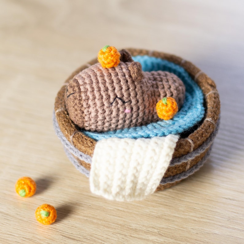 May include: A crocheted brown capybara with orange accents is relaxing in a blue and brown crocheted tub. The tub has a white crocheted towel hanging over the side.