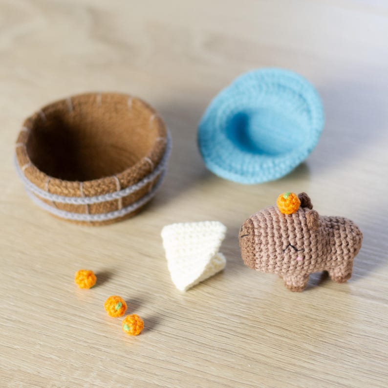 May include: A brown crocheted capybara with an orange crocheted pumpkin on its head. There are three small orange crocheted pumpkins on a wooden surface. There is a brown crocheted basket and a light blue crocheted hat on the surface.