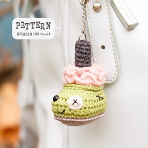 May include: A handmade crochet keychain in the shape of a green bear-like character with a pink top and a dark gray stem. The keychain is attached to a silver chain and is hanging from a white bag. The text "PATTERN ENGLISH (US terms)" is in the upper left corner.