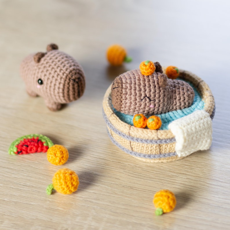 May include: A brown crocheted capybara relaxes in a wooden tub filled with water and orange crocheted fruit. The capybara is surrounded by more orange crocheted fruit.