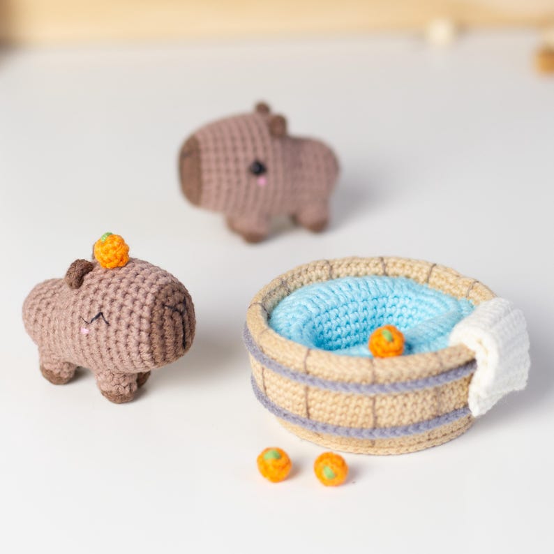 May include: Three brown crocheted capybara figurines with orange accents, one sitting in a crocheted wooden hot tub with blue water and a white towel. Three orange spheres are scattered on the white surface.