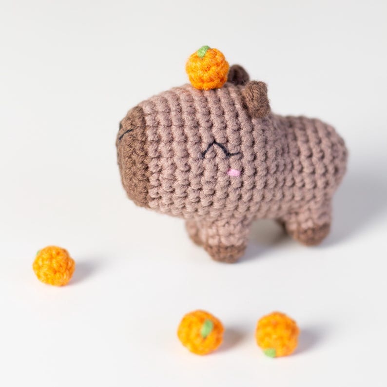 May include: A brown crocheted capybara with a small orange crocheted pumpkin on its head. Three additional orange crocheted pumpkins are on a white surface.