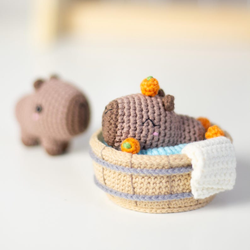 May include: A brown crocheted capybara plush toy is relaxing in a small crocheted bathtub with orange and green accents. The capybara has a happy expression and is surrounded by small orange and green balls.