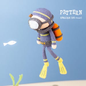 Puede incluir: Muñeco de buzo tejido a ganchillo en azul, amarillo y naranja. El muñeco tiene una máscara de buceo, un tanque de buceo y aletas. El texto "PATTERN ENGLISH (US terms" es visible. El fondo es azul claro.