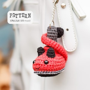 Może przedstawiać: Ręcznie robiony czerwony brelok do kluczy w kształcie smoka, z czarnymi akcentami i szarym ogonem. Brelok jest przymocowany do srebrnego łańcuszka. Widoczny jest tekst "PATTERN ENGLISH (US terms)".