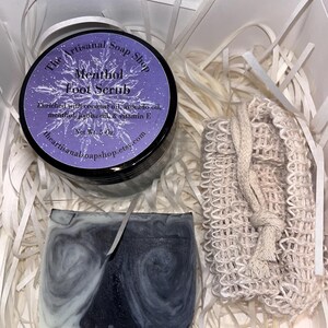 May include: A gift box filled with white shredded paper contains a jar of menthol foot scrub, a bar of soap, and a white mesh bag. The jar is labeled "The Artisanal Soap Shop Menthol Foot Scrub" and lists the ingredients as coconut oil, avocado oil, menthol, jojoba oil, and vitamin E. The jar is 5 oz.