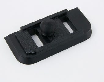 May include: A black, 3D-printed camera accessory. The rectangular base has three rectangular cutouts and a textured surface. A small, round knob is centered on the top. The accessory is designed to attach to a camera.