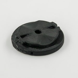 Op de afbeelding: Een zwart, cirkelvormig object van plastic, 3D-geprint, met een centraal gat en een reeks concentrische ringen.