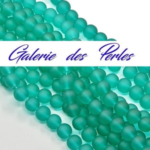 Peut inclure: Un brin de perles en verre mat vert turquoise. Les perles sont rondes et ont une finition lisse et givrée. Les perles sont disposées en rangée, avec le texte "Galerie des Perles" visible en arrière-plan.