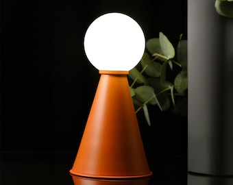 Può includere: Una lampada da tavolo moderna con un globo bianco e una base conica in terracotta. La lampada si riflette su una superficie scura.