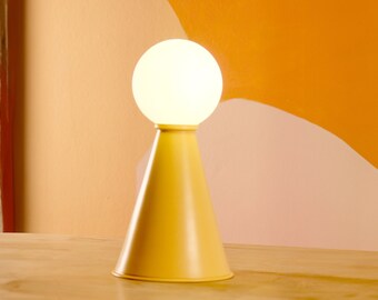 Può includere: Una lampada da tavolo conica gialla con un globo bianco. La lampada è su un tavolo di legno.