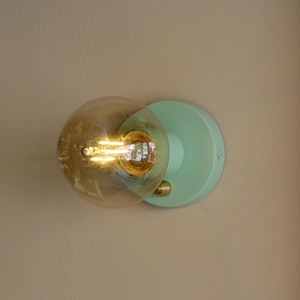 Op de afbeelding: Een lichtgroene wandlamp met een ronde, heldere glazen bolvormige lamp. De lamp heeft een gouden gloeidraad en is aan.