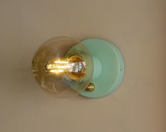 Può includere: Un'applique da parete verde chiaro con una lampadina rotonda in vetro trasparente. La lampadina ha un filamento dorato ed è accesa.