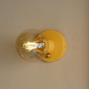 May include: A yellow wall sconce with a clear glass globe light bulb. The light bulb is turned on and emits a warm glow.