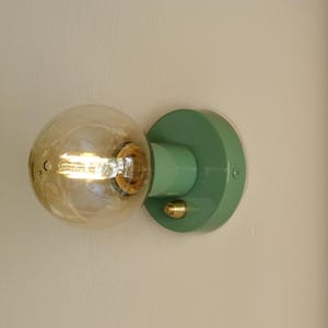 May include: A green wall-mounted light fixture with a round, frosted glass globe and a brass accent. The light is turned on and the bulb is visible.