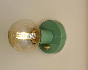 Può includere: Un apparecchio di illuminazione a parete verde con un globo di vetro smerigliato rotondo e un accento in ottone. La luce è accesa e la lampadina è visibile.