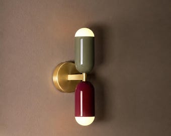 Può includere: Applique moderno con base in ottone e due lampade cilindriche. La lampada superiore è verde oliva, quella inferiore bordeaux. Entrambe hanno una luce bianca e brillante nella parte inferiore.