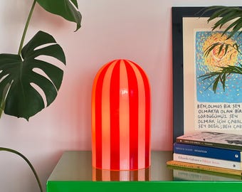 Può includere: Una lampada da tavolo a righe rosse e arancioni con la parte superiore arrotondata. La lampada è cilindrica e poggia su una base color oro. La lampada è su una superficie verde con una pianta e un'opera d'arte sullo sfondo.