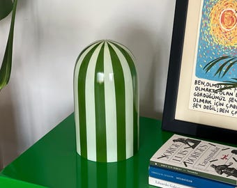 Può includere: Una campana di vetro decorativa a righe verdi e bianche. La campana è cilindrica con la parte superiore arrotondata ed è posta su una superficie verde. Un'opera d'arte incorniciata e una pila di libri sono sullo sfondo.