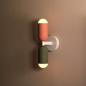 May include: A modern wall sconce with two light fixtures. The fixtures are cylindrical and made of ceramic. The top fixture is a light pink colour and the bottom fixture is a dark green colour. The sconce is mounted on a white circular base.