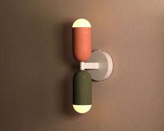 Può includere: Un'applique da parete moderna con due lampade. Le lampade sono cilindriche e in ceramica. La lampada superiore è di colore rosa chiaro e la lampada inferiore è di colore verde scuro. L'applique è montata su una base circolare bianca.