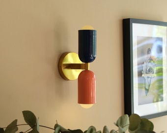 Può includere: Un'applique da parete in ottone con due punti luce. Il punto luce superiore è un cilindro blu scuro e il punto luce inferiore è un cilindro corallo.