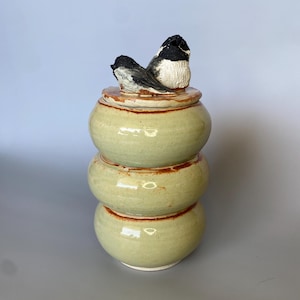 May include: A ceramic jar with a lid featuring two black and white birds. The jar is green with brown accents and has three stacked sections.