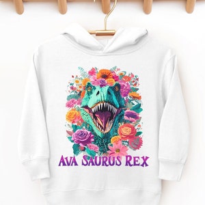 Toddler hooded sweatshirt in white with a graphic print of a roaring Tyrannosaurus rex and vibrant flowers around it. Below the dinosaur, text reads “Ava Saurus Rex”