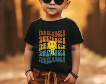 May include: A young child wearing a black t-shirt with a rainbow design and the text "THREE-NAGER" repeated multiple times. The child is wearing brown pants and black boots. The child is standing on a path in a forest.