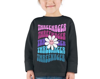 Puede incluir: Una sudadera negra de manga larga con un gráfico de margarita colorido y el texto "Threeenager" repetido en un patrón apilado.