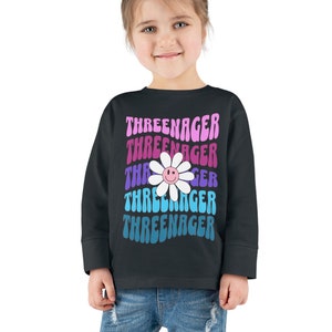 May include: A black long-sleeved sweatshirt with a colorful daisy graphic and the text "Threeenager" repeated in a stacked pattern.