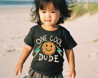 May include: A young child wearing a black t-shirt with the words "ONE COOL DUDE" and a smiling face graphic. The child is standing on a sandy beach, with the ocean and dunes in the background. The shirt has a retro design.