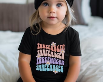 Puede incluir: Un niño pequeño con una camiseta negra con el texto "THREEENAGER" repetido cuatro veces en colores arcoíris con un pequeño gráfico de flores al lado de cada palabra. El niño lleva un turbante negro en la cabeza y tiene el pelo largo y rubio.