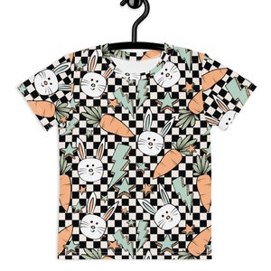 Puede incluir: Una camiseta de manga corta con un estampado repetido de caras de conejos blancos, zanahorias naranjas, rayos azul claro y estrellas sobre un fondo a cuadros blanco y negro. La camiseta está colgada de una percha negra.