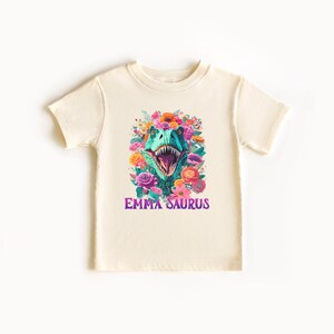 May include: A cream colored t-shirt with a green dinosaur head surrounded by colorful flowers. The text "EMMA SAURUS" is printed in purple below the dinosaur.