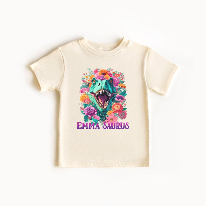 May include: A cream colored t-shirt with a green dinosaur head surrounded by colorful flowers. The text "EMMA SAURUS" is printed in purple below the dinosaur.