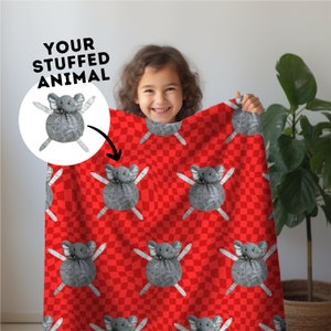 Puede incluir: Manta roja con un patrón repetido de juguetes de elefante de peluche grises. La manta tiene un diseño a cuadros. El texto "YOUR STUFFED ANIMAL" está en la esquina superior izquierda.